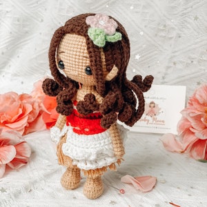 May include: A handmade crochet doll with brown hair, a pink and green flower, and a white and red dress. The doll has a tan face and limbs, and is posed against a backdrop of pink flowers.