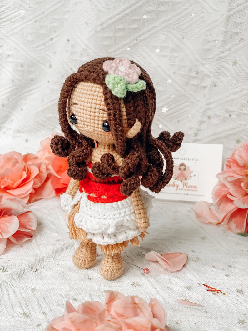 May include: A handmade crochet doll with brown hair, a pink and green flower, and a white and red dress. The doll has a tan face and limbs, and is posed against a backdrop of pink flowers.