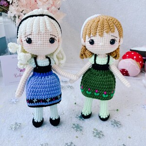 Puede incluir: Dos mu&ntilde;ecas de crochet vestidas con vestidos azules y verdes con detalles blancos. Las mu&ntilde;ecas tienen el pelo casta&ntilde;o y los ojos negros. Las mu&ntilde;ecas est&aacute;n de pie sobre una superficie blanca con copos de nieve plateados.