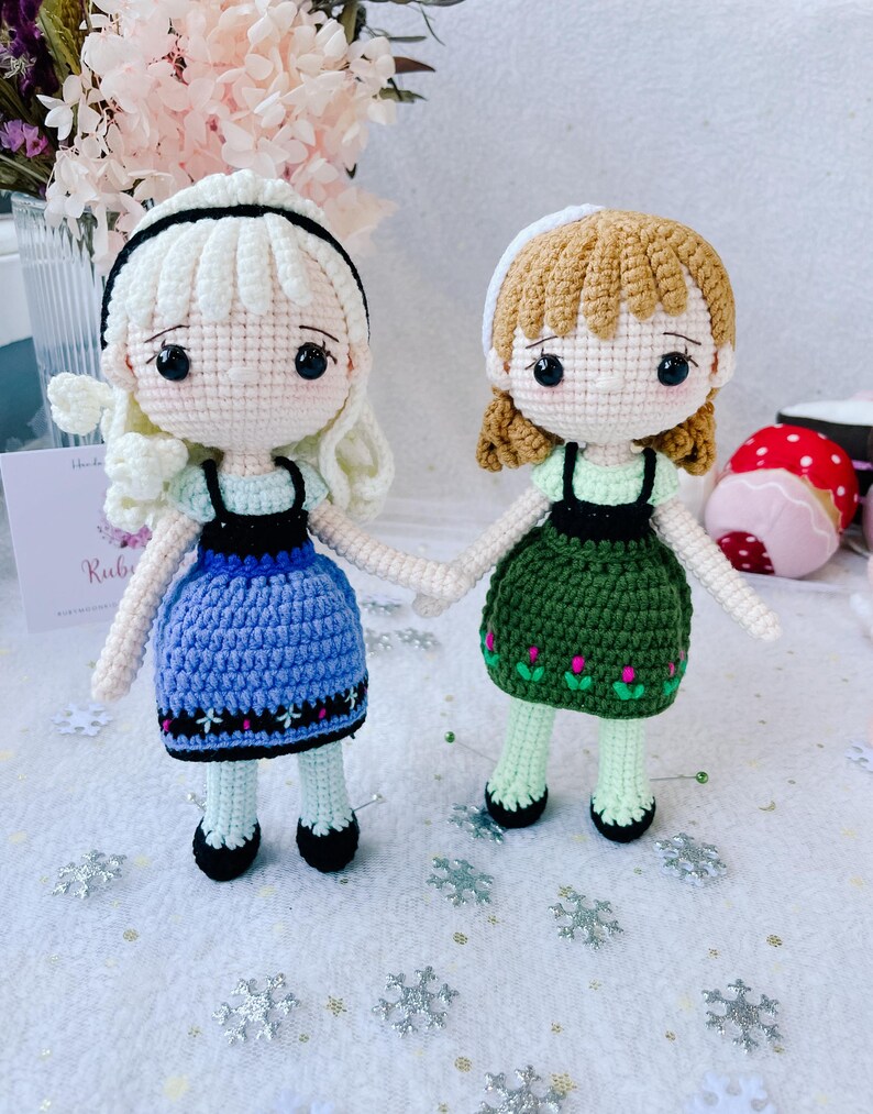 Puede incluir: Dos mu&ntilde;ecas de crochet vestidas con vestidos azules y verdes con detalles blancos. Las mu&ntilde;ecas tienen el pelo casta&ntilde;o y los ojos negros. Las mu&ntilde;ecas est&aacute;n de pie sobre una superficie blanca con copos de nieve plateados.