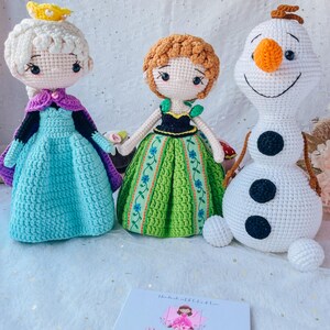 May include: Three crocheted dolls, Elsa, Anna, and Olaf from the movie Frozen. Elsa is wearing a light blue dress with a purple cape and a gold crown. Anna is wearing a green dress with a black bodice. Olaf is a white snowman with a carrot nose and black eyes.