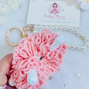 Puede incluir: Llavero de peluche rosa con detalles blancos, que se asemeja a una oveja o un cordero. El llavero est&aacute; unido a un anillo de llave dorado.