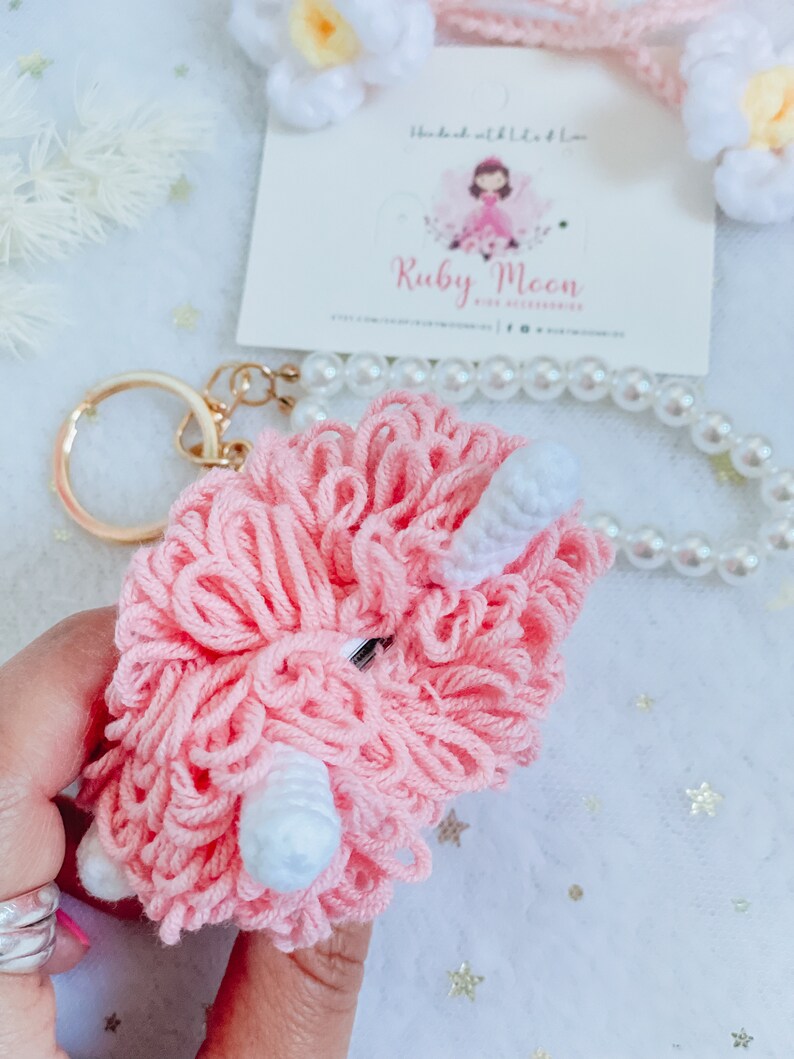 Puede incluir: Llavero de peluche rosa con detalles blancos, que se asemeja a una oveja o un cordero. El llavero est&aacute; unido a un anillo de llave dorado.