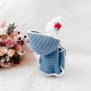 May include: A handmade crochet doll wearing a blue hooded coat with white trim and a white hat adorned with a red and blue flower. The doll is set against a soft, textured background, showcasing the intricate details of the crochet work.