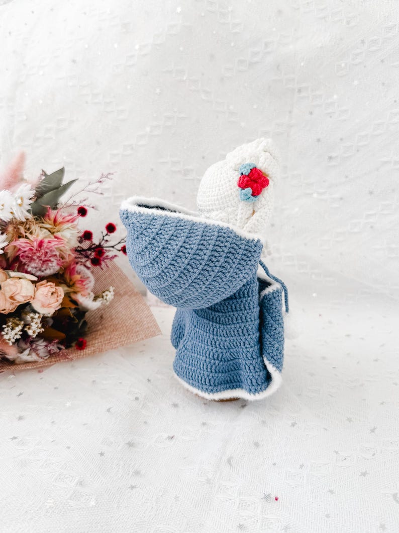 May include: A handmade crochet doll wearing a blue hooded coat with white trim and a white hat adorned with a red and blue flower. The doll is set against a soft, textured background, showcasing the intricate details of the crochet work.