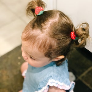 K&ouml;nnte beinhalten: Ein kleines Kind mit braunen Haaren tr&auml;gt ein hellblaues Kleid mit wei&szlig;en und rosa Verzierungen. Das Kind hat zwei haarf&ouml;rmige Haarklammern in Form von Erdbeeren im Haar.