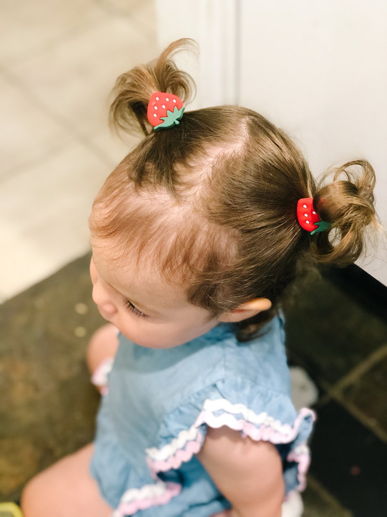 K&ouml;nnte beinhalten: Ein kleines Kind mit braunen Haaren tr&auml;gt ein hellblaues Kleid mit wei&szlig;en und rosa Verzierungen. Das Kind hat zwei haarf&ouml;rmige Haarklammern in Form von Erdbeeren im Haar.