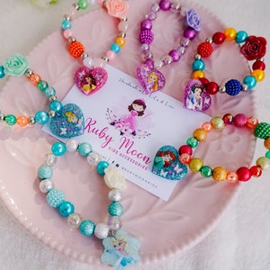 May include: A collection of colorful beaded bracelets with princess-themed charms. Each bracelet features a different princess character, including Cinderella, Ariel, and Elsa. The bracelets are displayed on a pink plate with a business card that reads "Ruby Moon Kids Accessories".