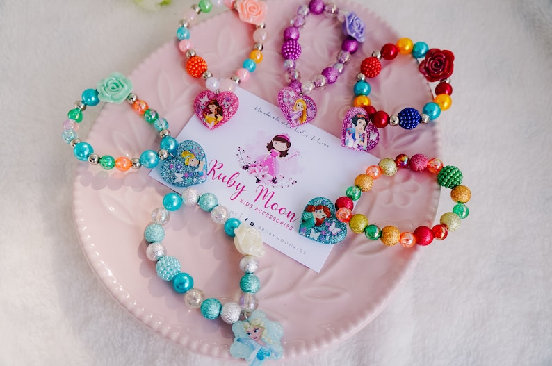 May include: A collection of colorful beaded bracelets with princess-themed charms. Each bracelet features a different princess character, including Cinderella, Ariel, and Elsa. The bracelets are displayed on a pink plate with a business card that reads "Ruby Moon Kids Accessories".