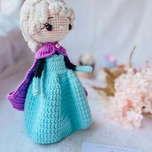 May include: A crocheted doll wearing a blue dress, purple cape, and a yellow crown. The doll has white hair and a pink face. The doll is handmade and is a kids accessory.
