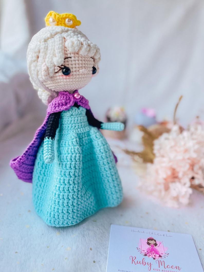 May include: A crocheted doll wearing a blue dress, purple cape, and a yellow crown. The doll has white hair and a pink face. The doll is handmade and is a kids accessory.