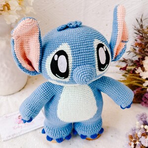 Op de afbeelding: Een blauw gehaakt Stitch knuffel met roze oren, witte ogen en een lichtblauwe buik. Het speelgoed staat op een wit oppervlak.