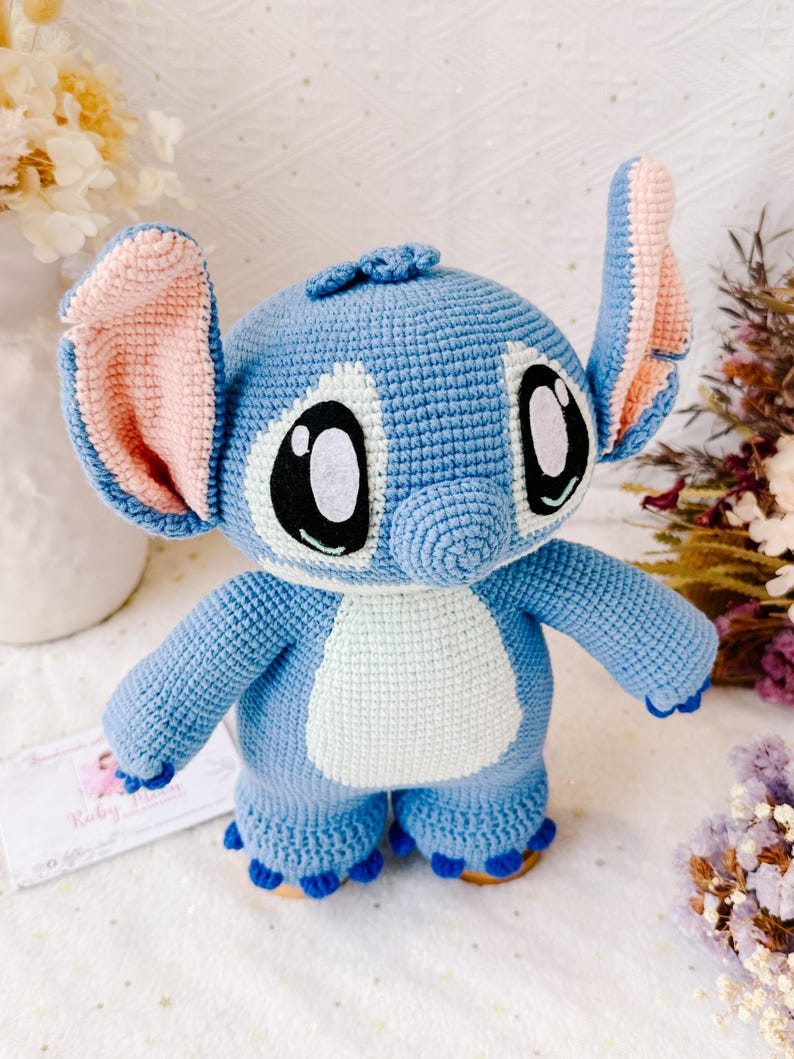 Op de afbeelding: Een blauw gehaakt Stitch knuffel met roze oren, witte ogen en een lichtblauwe buik. Het speelgoed staat op een wit oppervlak.