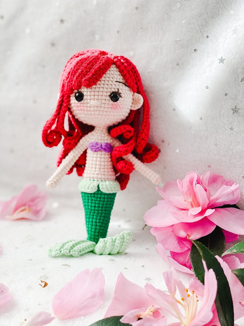 May include: A crocheted mermaid doll with red hair, a purple seashell bra, and a green tail. The doll is sitting on a white surface with pink flower petals.
