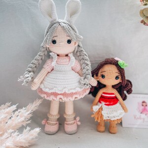 May include: Two handmade crochet dolls. One doll is a girl with gray hair, bunny ears, and a pink and white dress. The other doll is a girl with brown hair and a red and white dress.