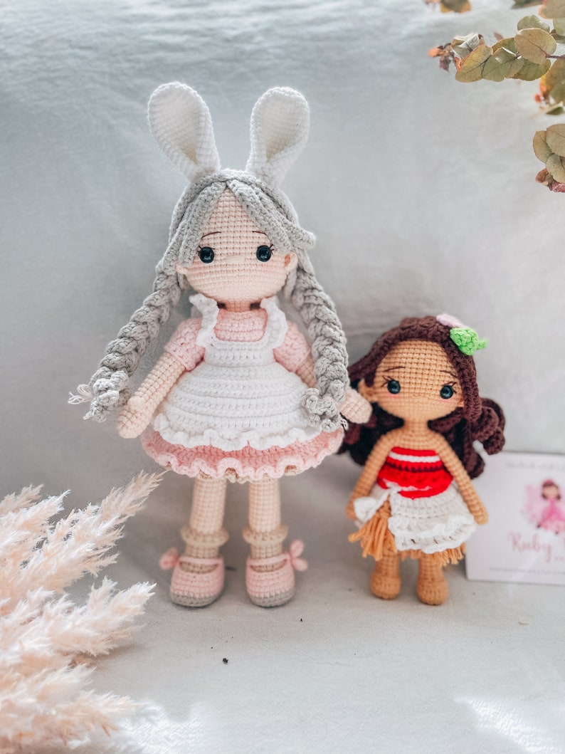 May include: Two handmade crochet dolls. One doll is a girl with gray hair, bunny ears, and a pink and white dress. The other doll is a girl with brown hair and a red and white dress.