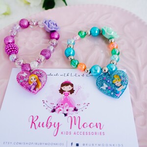May include: Two colorful beaded bracelets with heart-shaped charms featuring Disney princesses Rapunzel and Cinderella. The bracelets are made with iridescent beads and glitter. The charms are pink and blue with glitter and the princesses are depicted in their signature outfits.