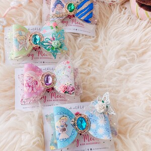 May include: Three handmade hair bows featuring Disney characters. The first bow is blue and gold with a green gem and a picture of Jasmine from Aladdin. The second bow is pink and green with a purple gem and a picture of Tinkerbell. The third bow is blue and white with a blue gem and a picture of Alice from Alice in Wonderland.