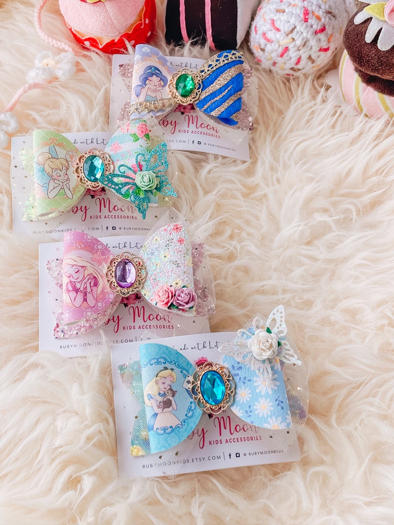 May include: Three handmade hair bows featuring Disney characters. The first bow is blue and gold with a green gem and a picture of Jasmine from Aladdin. The second bow is pink and green with a purple gem and a picture of Tinkerbell. The third bow is blue and white with a blue gem and a picture of Alice from Alice in Wonderland.