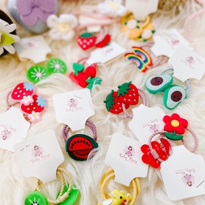 May include: Assortment of colourful hair ties with various fruit and animal designs. The hair ties are packaged on white cards with the text "Ruby Moon" and "Sweet Baby".