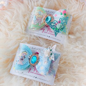 May include: Two handmade hair bows with a blue and green floral print. The bows feature a jeweled center and a butterfly accent. The text "by Moon Kids Accessories" is on the card.