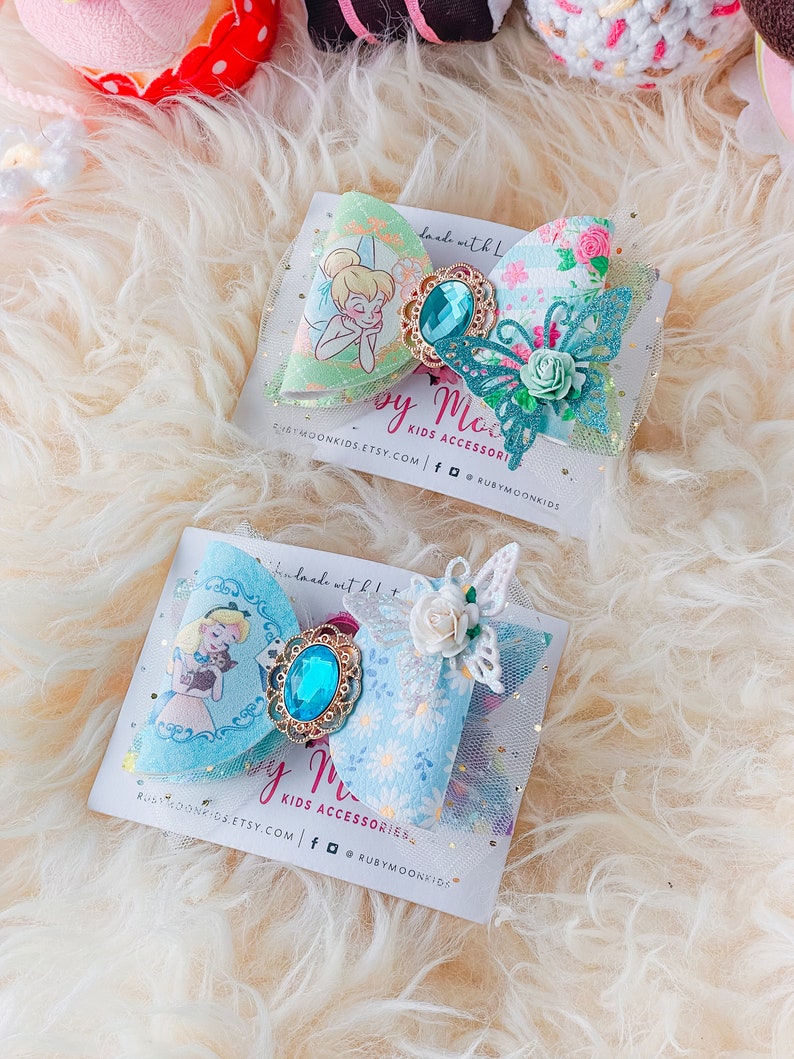May include: Two handmade hair bows with a blue and green floral print. The bows feature a jeweled center and a butterfly accent. The text "by Moon Kids Accessories" is on the card.