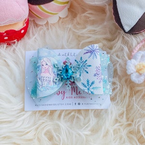May include: A light blue bow with a snowflake design and a sparkly blue gem. The bow features a printed image of Elsa from Frozen. The text "Ruby Moon Kids Accessories" is printed on the card below the bow.