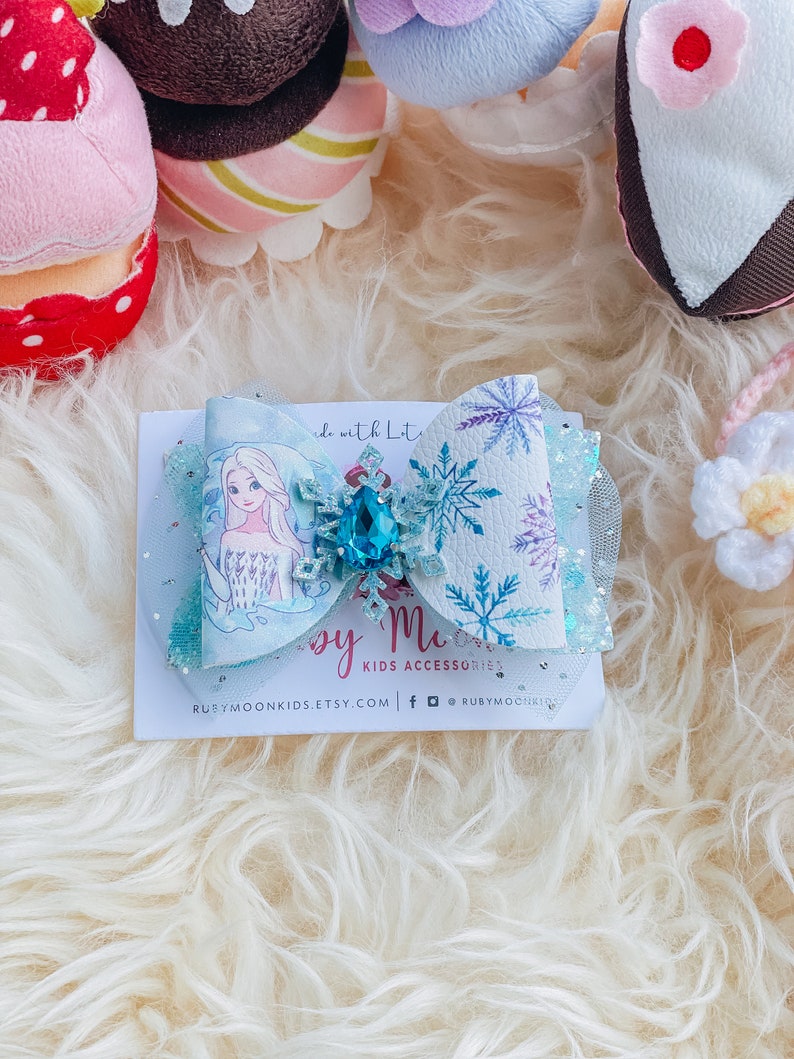 May include: A light blue bow with a snowflake design and a sparkly blue gem. The bow features a printed image of Elsa from Frozen. The text "Ruby Moon Kids Accessories" is printed on the card below the bow.