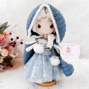 May include: A handmade crochet doll wearing a blue hooded cape with white trim and a light blue dress with snowflake accents. The doll has white hair and is standing on a wooden base. The doll is a handmade craft.