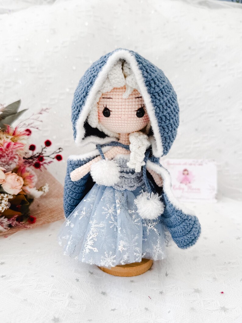 May include: A handmade crochet doll wearing a blue hooded cape with white trim and a light blue dress with snowflake accents. The doll has white hair and is standing on a wooden base. The doll is a handmade craft.