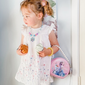 Puede incluir: Una ni&ntilde;a peque&ntilde;a con un vestido blanco con bordado floral, leggings a rayas y un collar de perlas con un dije de estrella azul y morado. Est&aacute; sosteniendo un juguete marr&oacute;n y uno verde y lleva un bolso rosa con el tema de Frozen con una correa de cadena plateada.