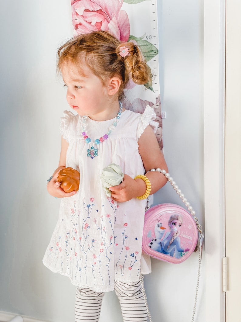 Puede incluir: Una ni&ntilde;a peque&ntilde;a con un vestido blanco con bordado floral, leggings a rayas y un collar de perlas con un dije de estrella azul y morado. Est&aacute; sosteniendo un juguete marr&oacute;n y uno verde y lleva un bolso rosa con el tema de Frozen con una correa de cadena plateada.