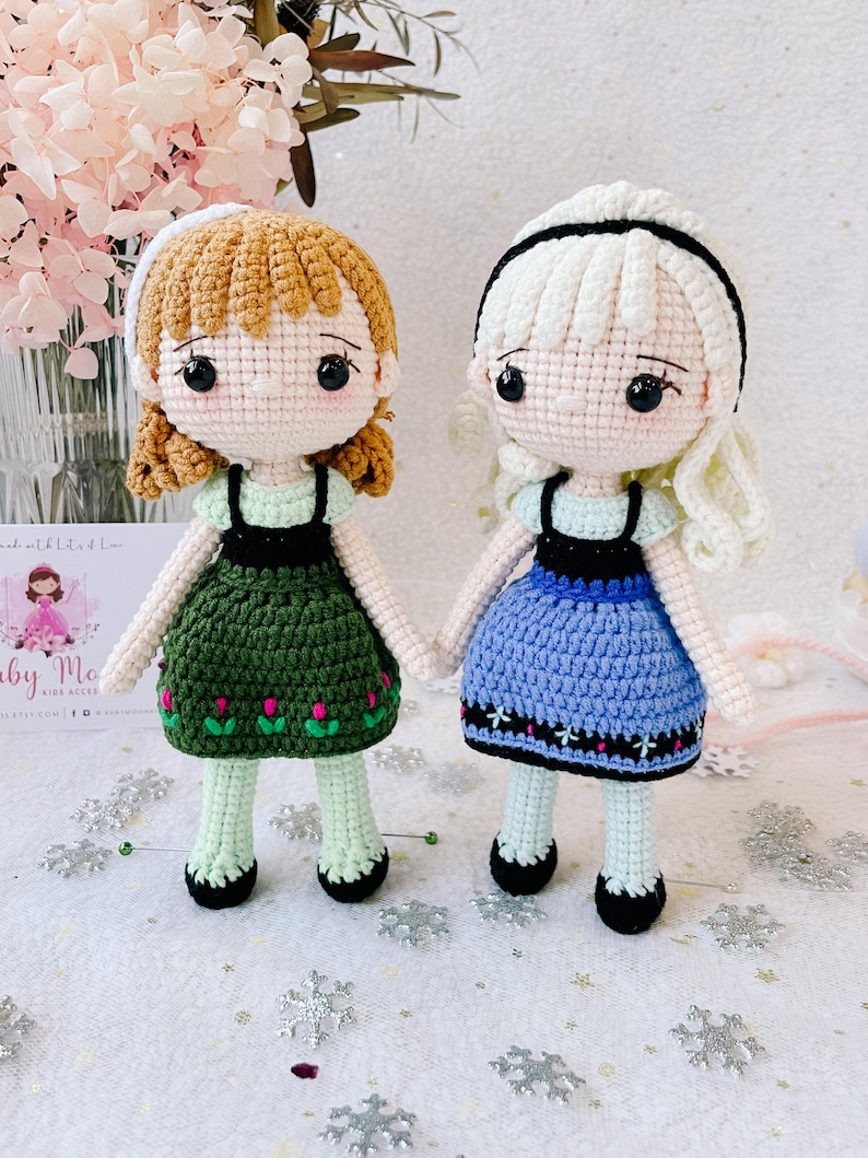 Puede incluir: Dos mu&ntilde;ecas de crochet, una con cabello casta&ntilde;o y un vestido verde, la otra con cabello rubio y un vestido azul. Ambas mu&ntilde;ecas tienen ojos negros y llevan zapatos negros.