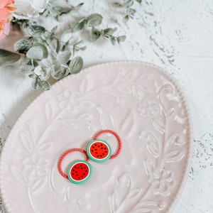 K&ouml;nnte beinhalten: Zwei rote und gr&uuml;ne Wassermelonen-Haargummis auf einem rosa Teller mit Blumenmuster.