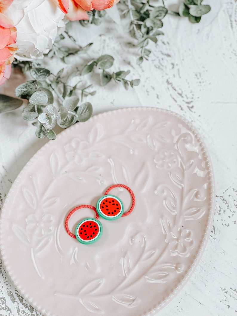 K&ouml;nnte beinhalten: Zwei rote und gr&uuml;ne Wassermelonen-Haargummis auf einem rosa Teller mit Blumenmuster.