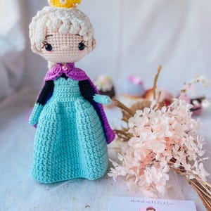 May include: A crocheted doll wearing a blue and purple dress with a yellow crown. The doll is standing on a white surface with pink flowers in the background. The text "Ruby Moon Kids Accessories" is visible on a white card.