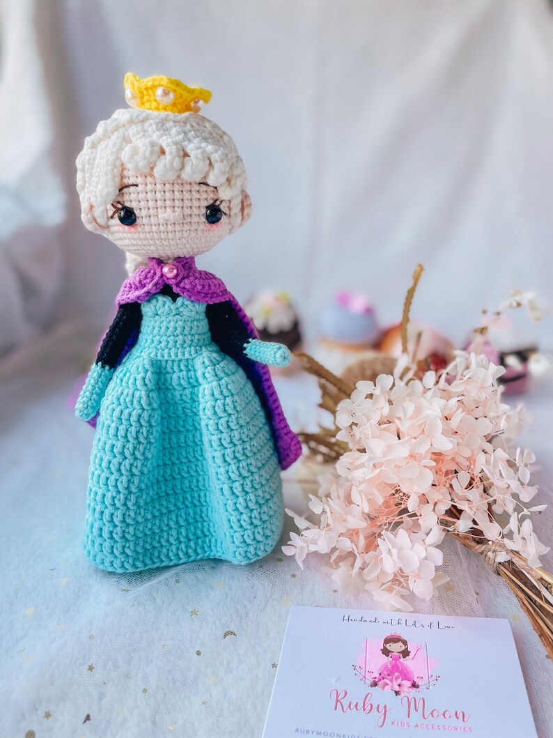 May include: A crocheted doll wearing a blue and purple dress with a yellow crown. The doll is standing on a white surface with pink flowers in the background. The text "Ruby Moon Kids Accessories" is visible on a white card.