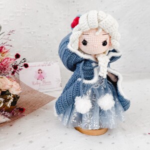 May include: A handmade crochet doll with a blue coat, white hair, and a snowflake-patterned skirt. The doll has a red and white hat and is displayed on a wooden base. The doll is a handcrafted item.