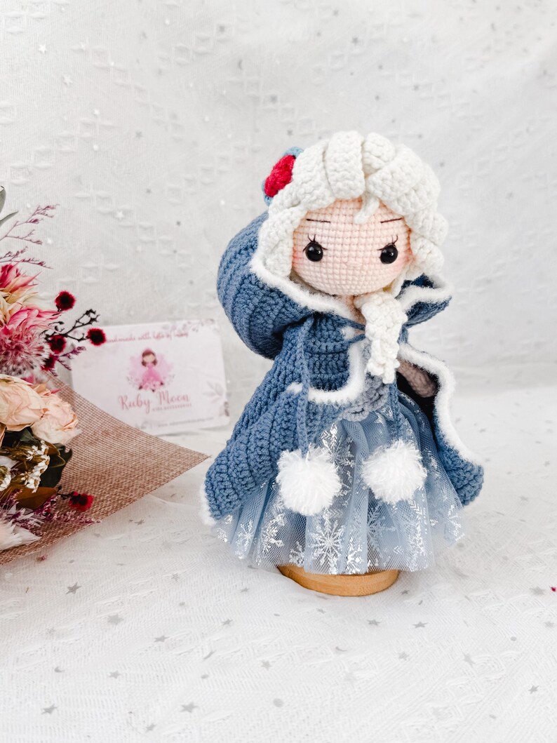 May include: A handmade crochet doll with a blue coat, white hair, and a snowflake-patterned skirt. The doll has a red and white hat and is displayed on a wooden base. The doll is a handcrafted item.