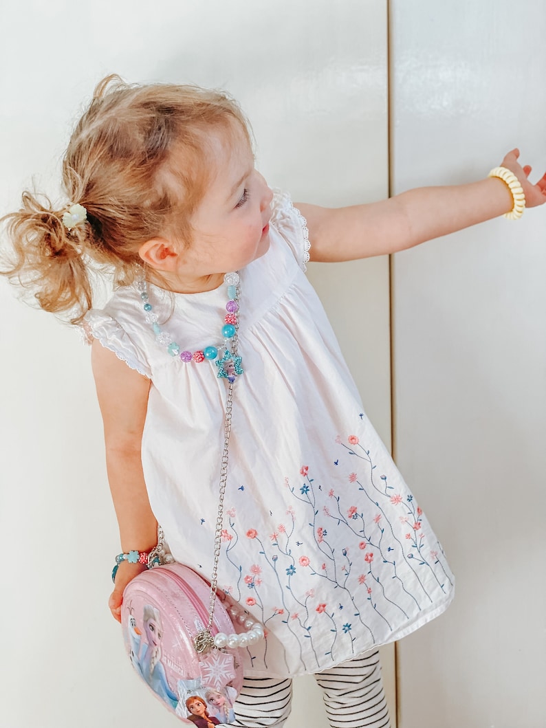 Puede incluir: Una ni&ntilde;a peque&ntilde;a con un vestido blanco con bordado floral, un collar de cuentas y un bolso rosa con un dise&ntilde;o de personaje de Frozen. Tambi&eacute;n lleva una pulsera amarilla y leggings a rayas.