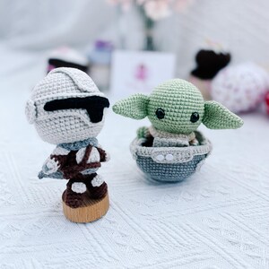 May include: Two crocheted figures: a Stormtrooper and a green alien character. The Stormtrooper is in a white and black helmet and brown uniform, mounted on a wooden base. The alien character is in a gray and blue pod.