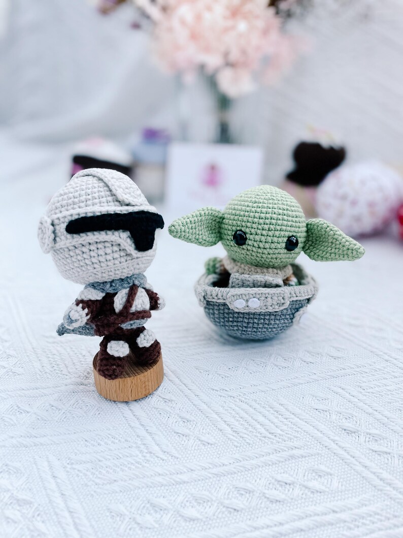 May include: Two crocheted figures: a Stormtrooper and a green alien character. The Stormtrooper is in a white and black helmet and brown uniform, mounted on a wooden base. The alien character is in a gray and blue pod.
