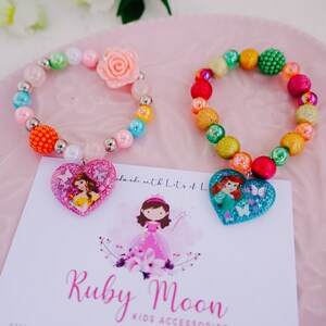 May include: Two colorful beaded bracelets with princess-themed heart charms. The bracelets are made with a variety of colorful beads, including glitter, iridescent, and solid colors. The charms feature images of Disney princesses, Belle and Ariel. The bracelets are displayed on a pink surface with a white card that reads "Ruby Moon Kids Accessories".