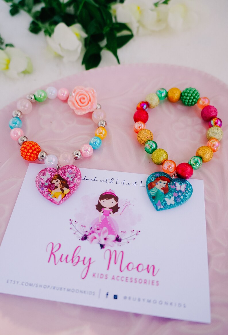 May include: Two colorful beaded bracelets with princess-themed heart charms. The bracelets are made with a variety of colorful beads, including glitter, iridescent, and solid colors. The charms feature images of Disney princesses, Belle and Ariel. The bracelets are displayed on a pink surface with a white card that reads "Ruby Moon Kids Accessories".