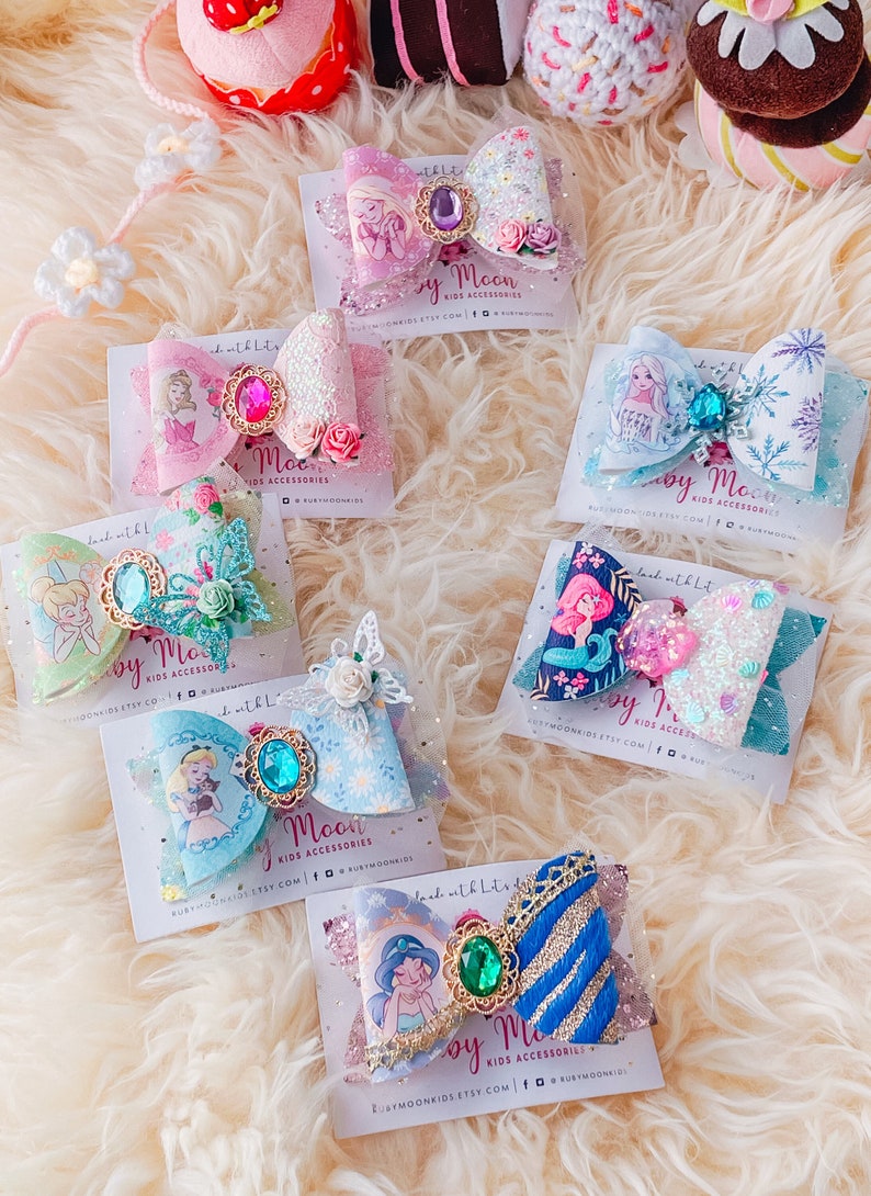 May include: A set of six colorful hair bows featuring various Disney princesses. Each bow is decorated with glitter, sequins, and a jeweled center. The bows are displayed on white cards with the text "Ruby Moon Kids Accessories" and their website address.