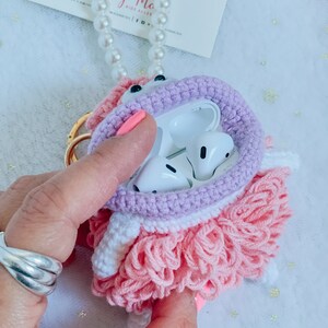 Puede incluir: Una funda para Airpods de crochet rosa y morado con una falda de volantes blanca y rosa. La funda est&aacute; abierta y muestra los Airpods blancos en el interior.