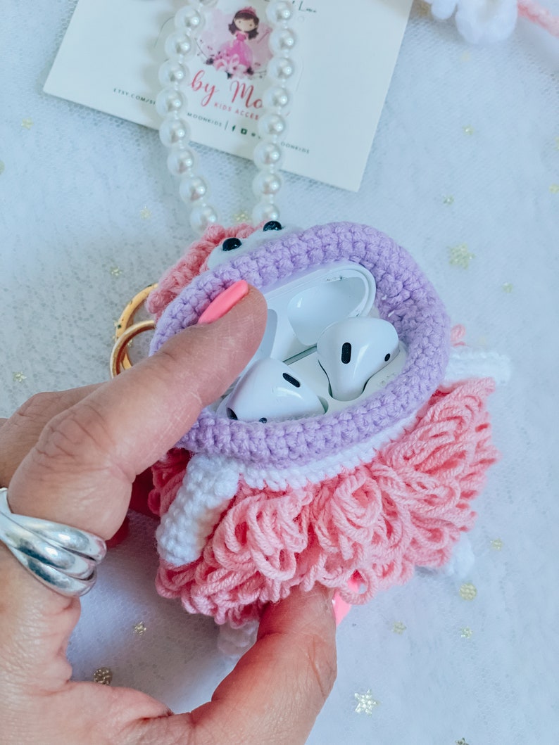 Puede incluir: Una funda para Airpods de crochet rosa y morado con una falda de volantes blanca y rosa. La funda est&aacute; abierta y muestra los Airpods blancos en el interior.