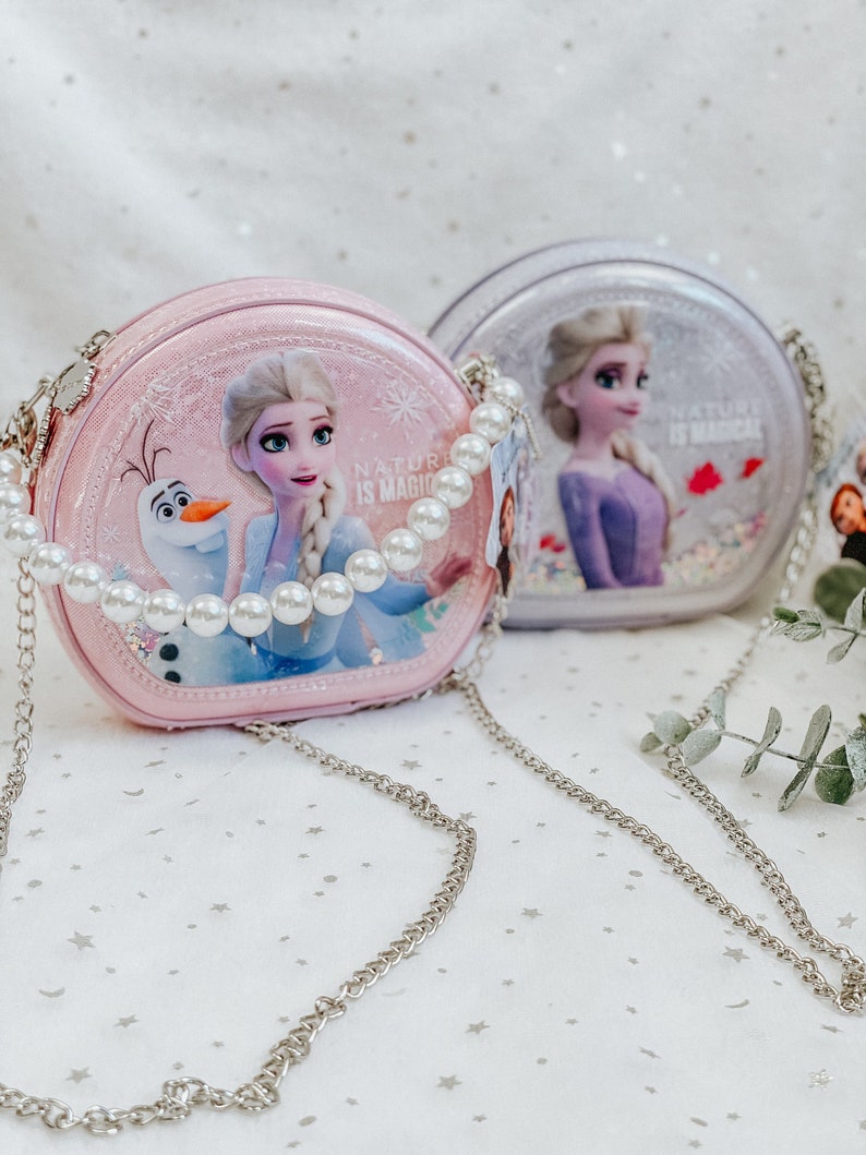 Puede incluir: Dos bolsos redondos de color rosa y morado con una correa de cadena plateada. Los bolsos tienen un acabado brillante e im&aacute;genes de Elsa y Anna de la pel&iacute;cula Frozen. El texto "Nature is Magic" est&aacute; impreso en los bolsos.