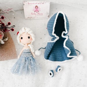 May include: A handmade crochet doll with a light blue dress and white hair, next to a blue hooded cape with white trim and two matching shoe covers. The doll has a sweet face. The Ruby Moon logo is visible on the packaging.