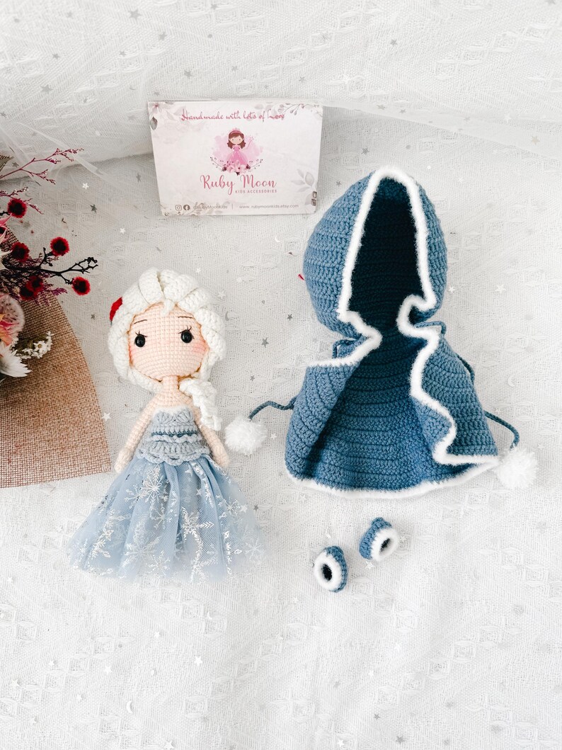 May include: A handmade crochet doll with a light blue dress and white hair, next to a blue hooded cape with white trim and two matching shoe covers. The doll has a sweet face. The Ruby Moon logo is visible on the packaging.